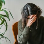 Unrecognizable young upset female millennial with dark hair in stylish sweater covering face with hand while siting on chair at home